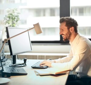 A man smiling while working at an office desk with a computer and natural daylight streaming in through large windows.